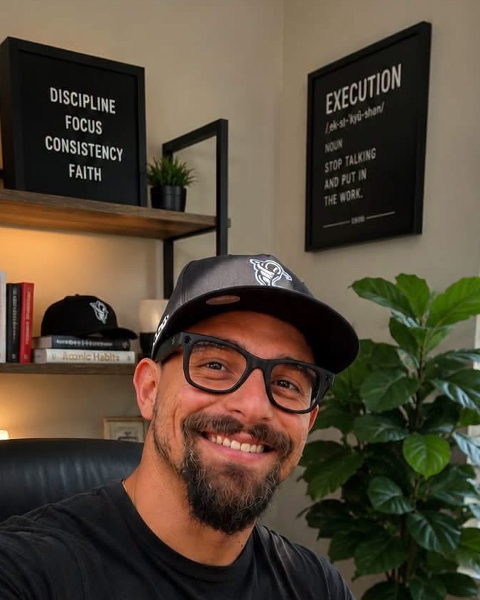 Angel Pichardo smiling at his desk. Framed prints behind him read Discipline, Focus, Consistency, Faith, and Execution: stop talking and put in the work.